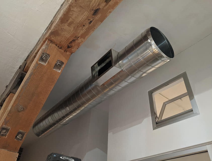 Round supply duct mounted beneath exposed beam